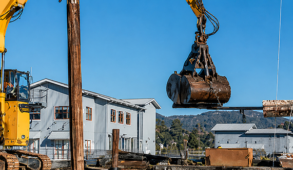 excavator-grapple-lifting-metal-tank-industrial-site Yellow excavator with a grapple attachment lifting a large metal tank at an industrial site