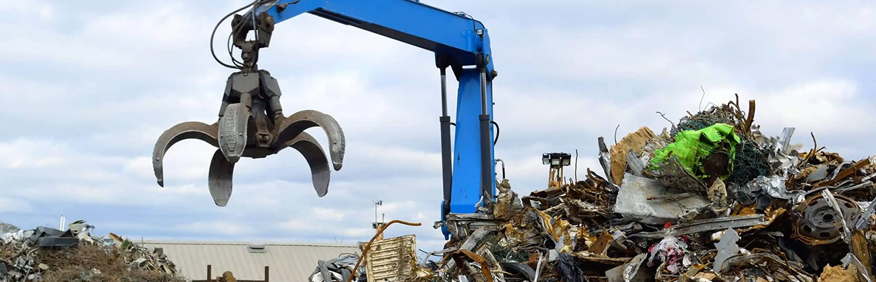 grapple-crane-scrap-metal-pile-wide-banner Blue grapple crane with an open orange peel grabber beside a large pile of scrap metal